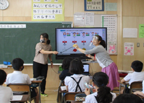 教室前方に置かれたモニターに映し出されたイラストを指示棒で指している黄土色の服を着た女性と、フェイスガードをつけたピンク色のスカートを履いた女性が左側を向いており、席についた生徒達を後方から写した写真